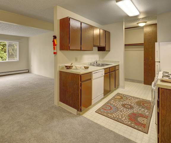 A view of the empty carpeted living room with a window and the kitchen with white appliances and wooden large cabinets at Taiga Apartment Homes. 