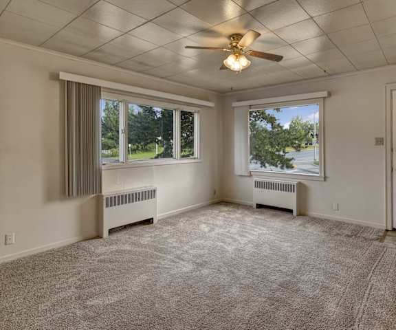 An empty living room with carpeting, two large windows with curtains, overhead lighting with a fan, and the front door at the Alaskan Apartment Homes. 