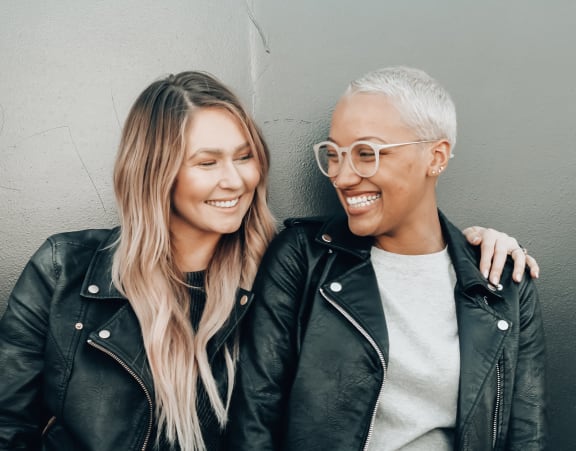 two women wearing leather jackets laughing and leaning against a wall