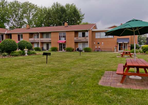A green umbrella is on a picnic table in front of a brick apartment building.
