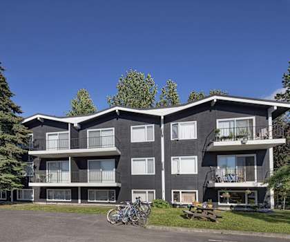 The black modern apartment buildings surrounded by the trees with grass and a picnic table in the front at the Alaskan Apartment Homes. 