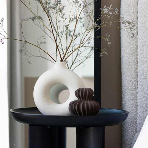 A white vase with a green circle inside sits on a black table next to a white pillow.
