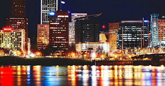 A city skyline at night with buildings illuminated and reflected in the water.