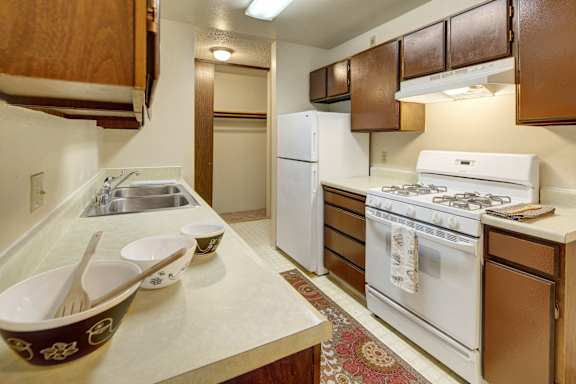A spacious/cozy kitchen with white appliances, kitchen utensils, overhead lighting, light countertops with lots of room, utensils, and large medium wooden cabinets at Taiga Apartment Homes.