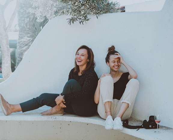 Two women sitting on a white wall outside.