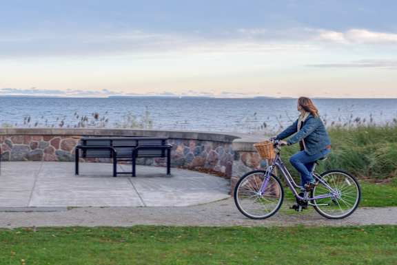 A woman rides a bicycle past a bench by the water.