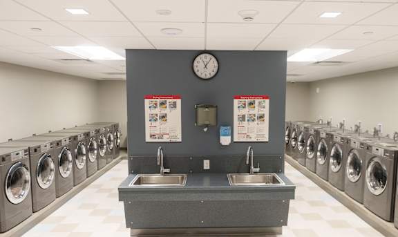 A laundromat with a clock on the wall and rows of washing machines at Highpoint Towers Apartments, Erie
