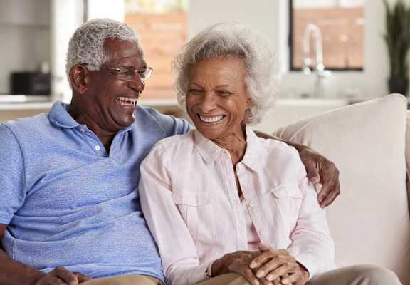 An older couple is sitting on a couch, smiling and laughing together.