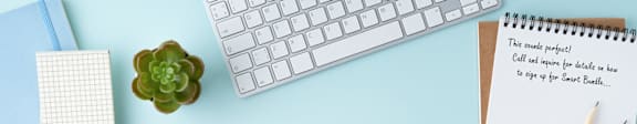 A white keyboard, a green plant, a notebook, and a pen are on a light blue surface.