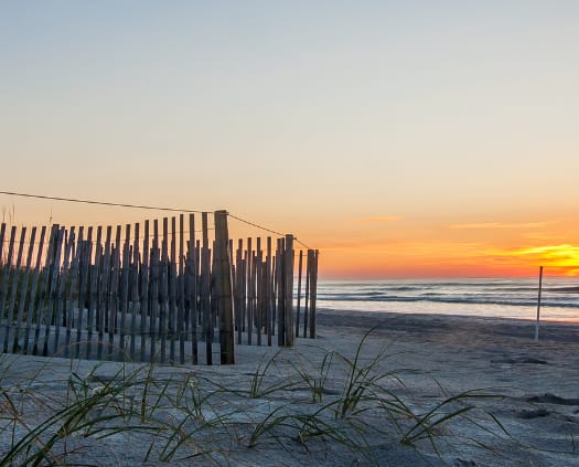 A wooden fence stands on a sandy beach with grass in the foreground.