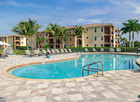 A swimming pool surrounded by a brick patio and palm trees in front of apartment buildings.