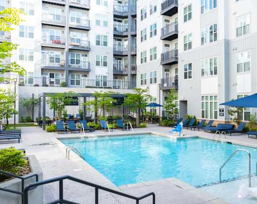 A swimming pool surrounded by lounge chairs and umbrellas in front of apartment buildings.