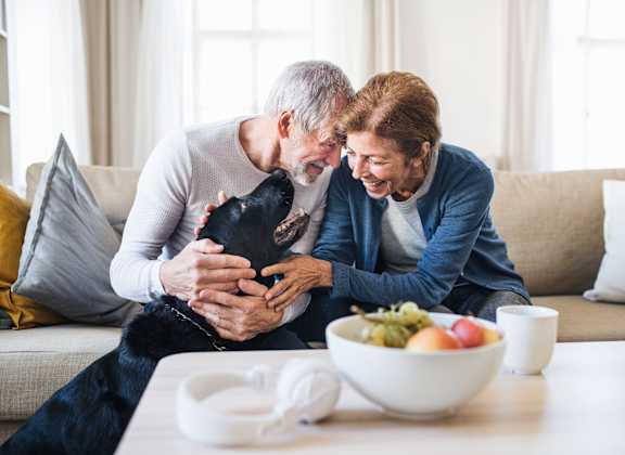 An elderly couple is sitting on a couch with a black dog between them.