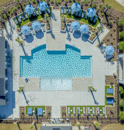 An aerial view of a swimming pool surrounded by a grassy area and several umbrellas. at Evolve St. James Apartment Homes Apartments, Southport, NC, 28461