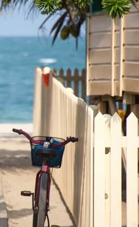 A bicycle with a basket is parked next to a white picket fence on a sunny day.