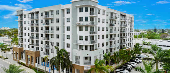 A large white building with a brown roof and balconies is surrounded by palm trees.