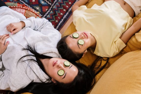 two women laying on a bed with slices of cucumber on their faces