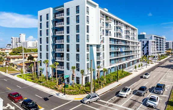 A modern white apartment building is situated on a street corner with cars parked and driving by.
