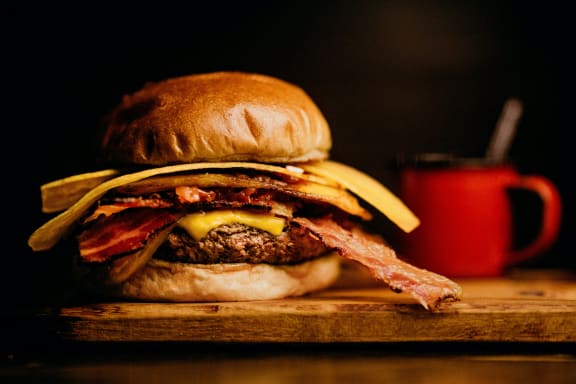 a hamburger with bacon and cheese on a wooden cutting board next to a cup