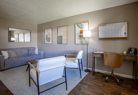 A living room with a grey couch, a white chair, a desk with a chair, a lamp, a calendar, and a mirror.