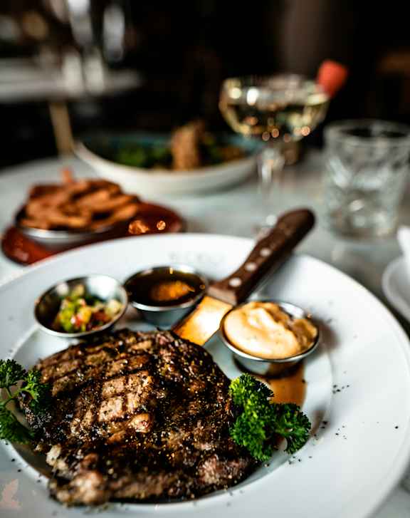 A steak is served on a white plate with a side of sauce and a piece of broccoli.
