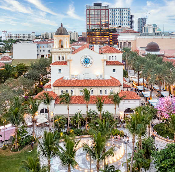 A white building with a clock tower is surrounded by palm trees.