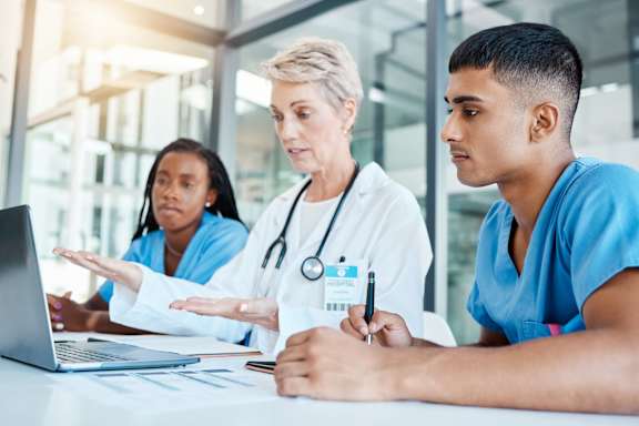 Three medical professionals are gathered around a laptop, discussing something important.