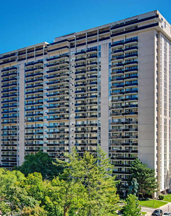 A tall building with many windows is surrounded by trees and has a parking lot in front.