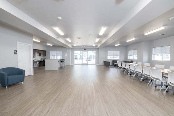 A room with white walls and wooden flooring has a long table with chairs and a speaker.