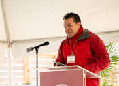 Thumbnail 17 of 24 - A man in a red jacket is giving a speech at a podium with a sign that says "BRIDGE Housing".