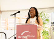 Thumbnail 16 of 24 - A woman is giving a speech at a podium with the words "BRIDGE Housing" on it.