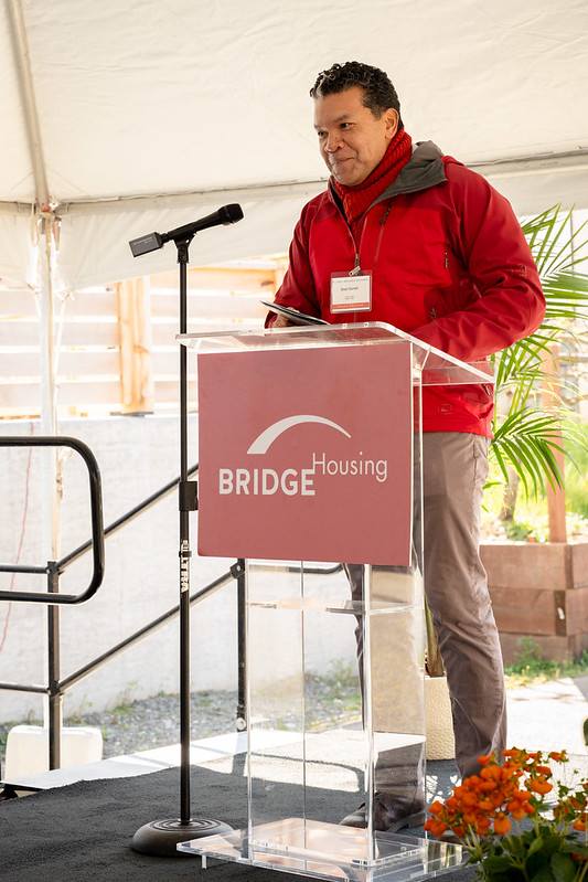 A man in a red jacket is giving a speech at a podium with a sign that says "BRIDGE Housing".