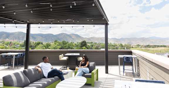 A man and a woman are sitting on a couch on a patio with mountains in the background.