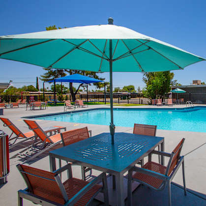 A table and chairs are set up under a blue umbrella by a pool.