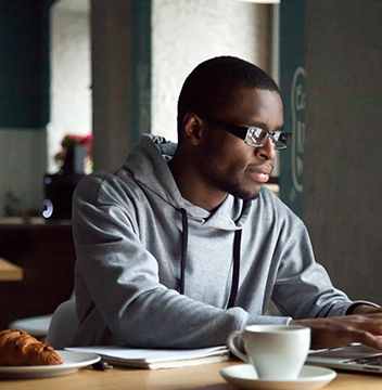 A man in a grey hoodie is using a laptop at a table with a plate of food and a cup of coffee.