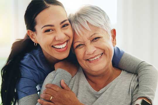A woman with grey hair is hugging a younger woman with dark hair.