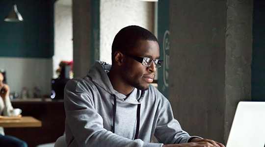 A man in a grey hoodie is using a laptop at a table with a plate of food and a cup of coffee.