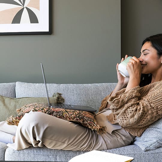 a woman sitting on a couch drinking from a mug and looking at her laptop