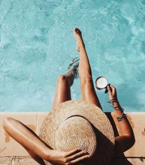 a woman laying on a pool side with a hat on and a clock in her hand