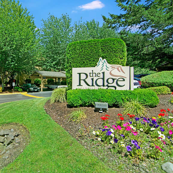 The exterior sign with trees and surrounded by greenery and flowers with the buildings and the parking lot in the background of the Ridge and Shores Apartment Homes. 