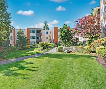 The beautiful exterior of the white, tan, and orange apartments and the green space with large swathes of grass, trees, and shrubs at Watercrest Apartment Homes. 