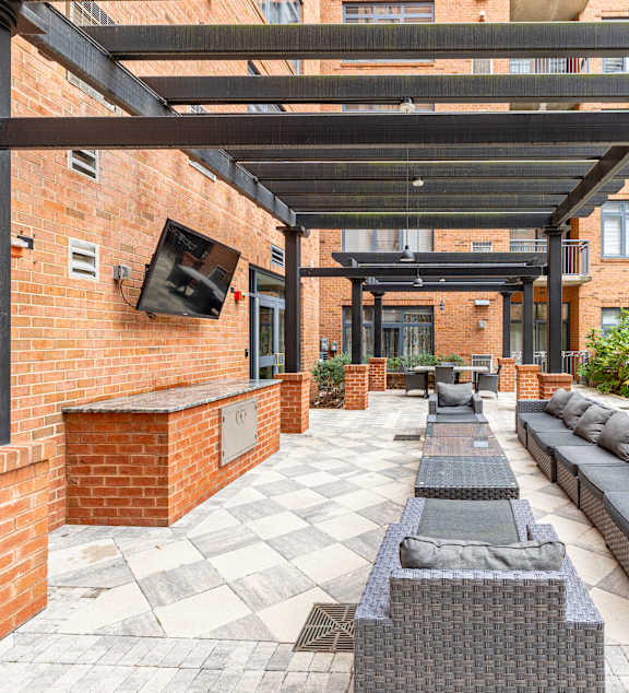 Outdoor courtyard patio featuring a black pergola, brick outdoor kitchen bar with grill, TV, and cushioned lounge seating at The Barton at Woodley in Washington DC