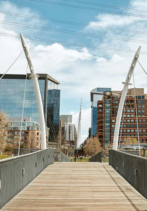 A bridge with a wooden walkway and metal railings spans a body of water with buildings in the background.
