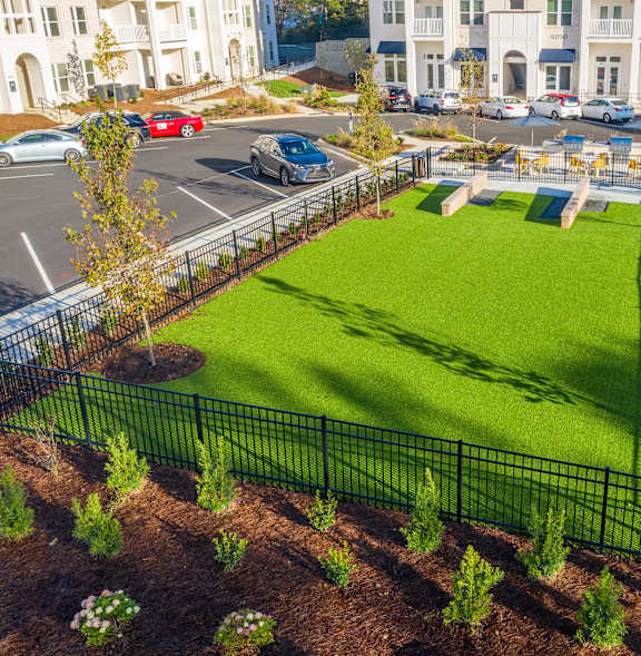 A green lawn surrounded by a black fence with trees and a parking lot in the background.
