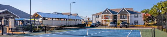 A tennis court with a blue surface and white lines, surrounded by a fence and a building in the background at Reserve of Bossier City Apartment Homes in Bossier City, LA 71111