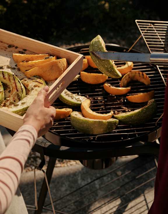 A person grilling slices of zucchini and pumpkin on a barbecue.