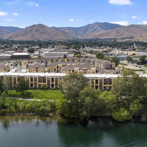 The arial view of the Riverside 9 Apartment Homes community, complete with a view of the mountains in the background, cityscape, and lakefront lined with trees. 