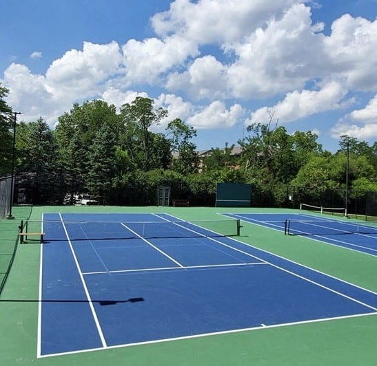A tennis court surrounded by trees under a blue sky with clouds. at Indian Creek Apartments, Cincinnati, OH, 45236