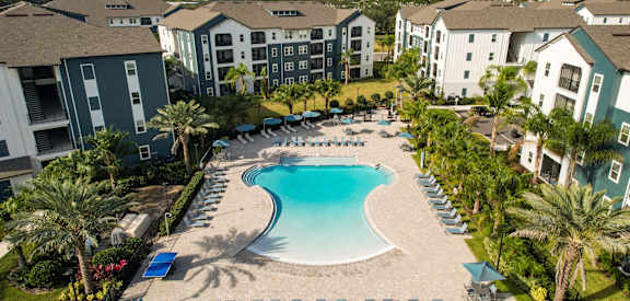 A swimming pool surrounded by palm trees and lounge chairs in front of apartment buildings.