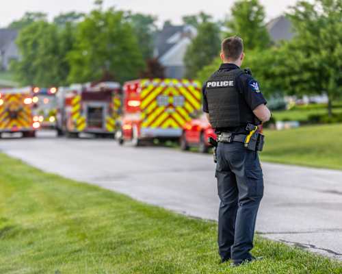 A police officer stands on the grass watching a line of fire trucks.